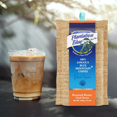 Plantation Blue 12 oz dark whole-bean Jamaica Blue Mountain coffee package next to a glass of iced milk coffee