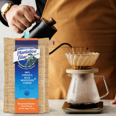 Plantation Blue 12 oz dark whole-bean Jamaica Blue Mountain coffee beside a hand pouring from a black kettle into a filter brewer