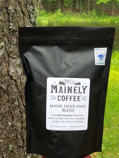 Black Maine Highlands bag propped beside a tree trunk in a green woodland