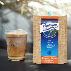 Plantation Blue 8 oz dark whole-bean Jamaica Blue Mountain coffee package next to a glass of iced milk coffee