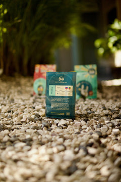 Back of a teal Tèt āndi Laye bag standing on gravel with other packages behind it