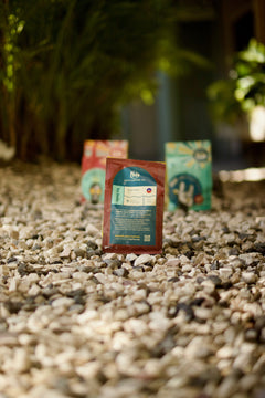 Back-label view of a red Kafe Dondon pouch on gravel with blurred bags behind
