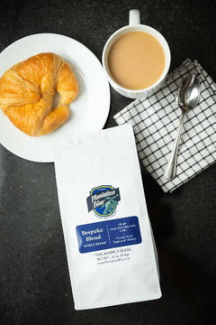 Bespoke whole-bean coffee bag beside a croissant and cup on a dark breakfast surface