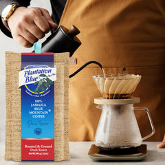 Plantation Blue 12 oz dark ground Jamaica Blue Mountain coffee beside a hand pouring from a black kettle into a filter brewer